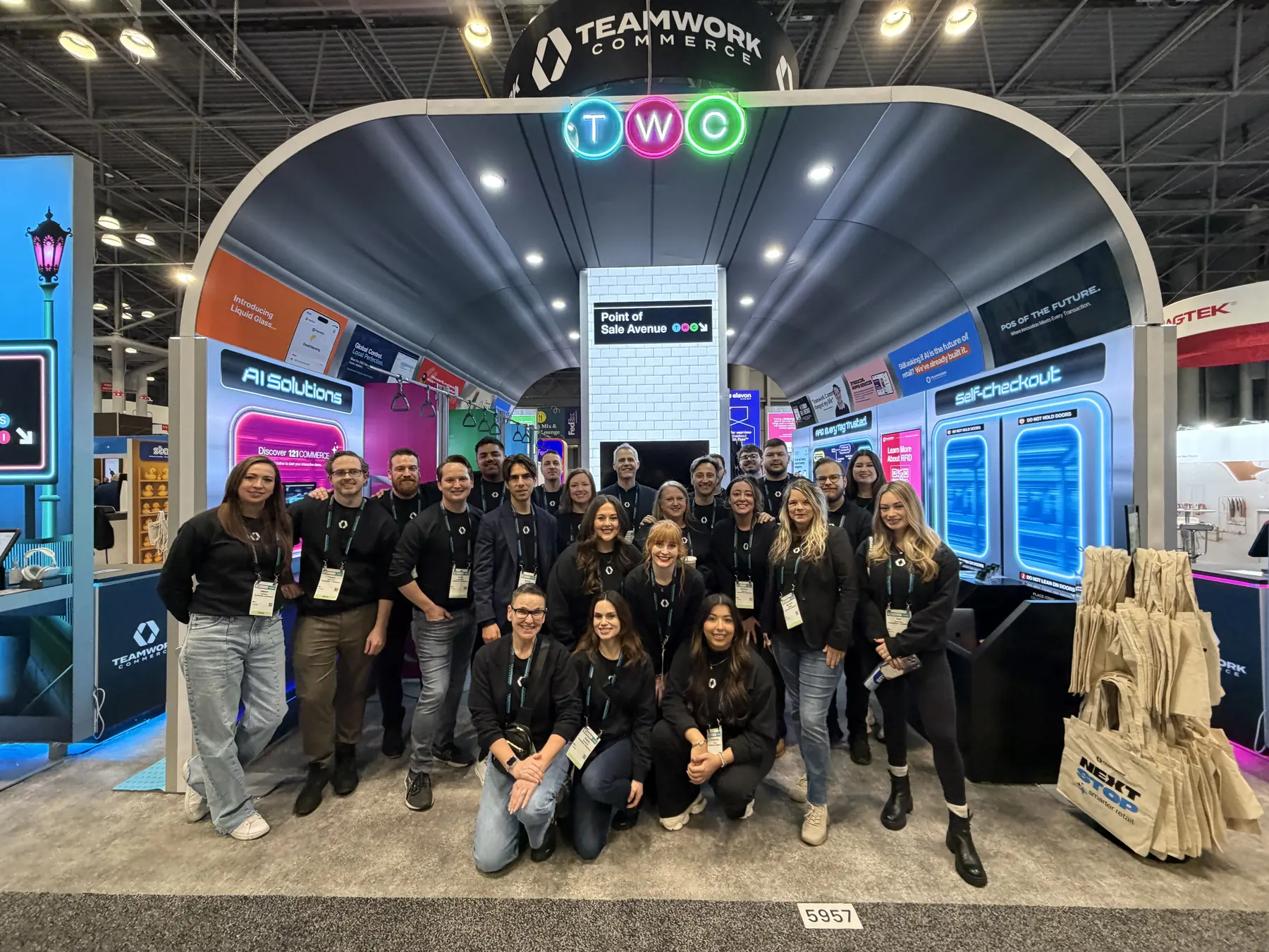The Teamwork Commerce team in front of their booth on the show floor, with the Point of Sale Avenue arch, AI Solutions and self-checkout displays behind them